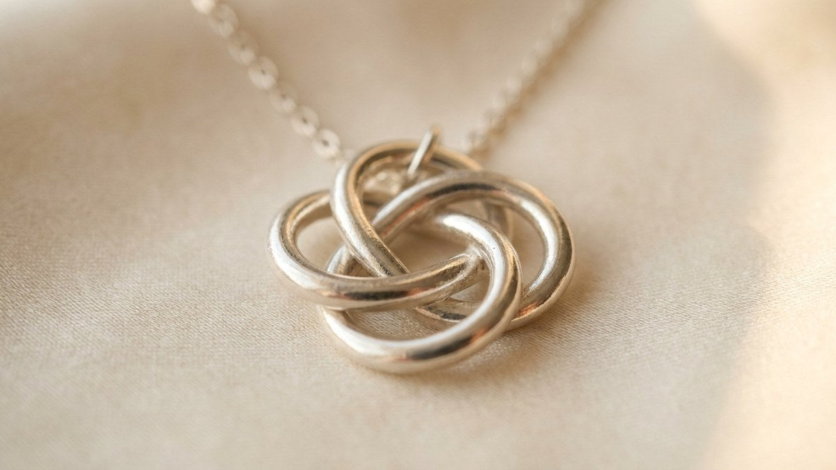 Colgante de plata con nudo de bruja de cuatro brazos entrelazados sobre seda crema, símbolo de protección.