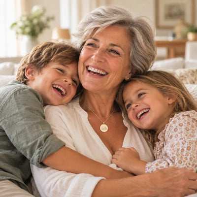 Abuela riendo abrazada por sus dos nietos en el sofá; lleva un colgante redondo de oro grabado con las siluetas de sus nietos