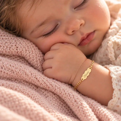 Bebé dormido sobre una manta de punto rosa con una pulsera de oro personalizada con su nombre grabado