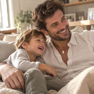 Padre sonriendo en el sofá con su hijo pequeño abrazado; lleva una cadena de plata con una placa rectangular personalizada